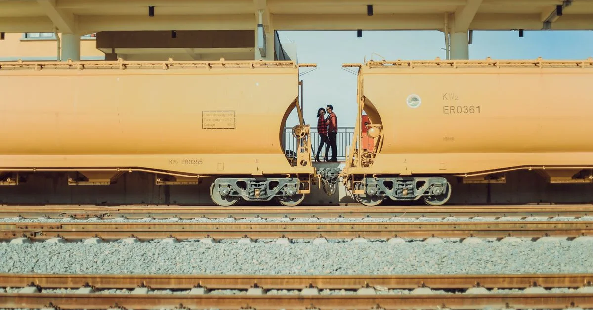 Deux individus posent entre des trains de marchandises connectés dans une gare ensoleillée.