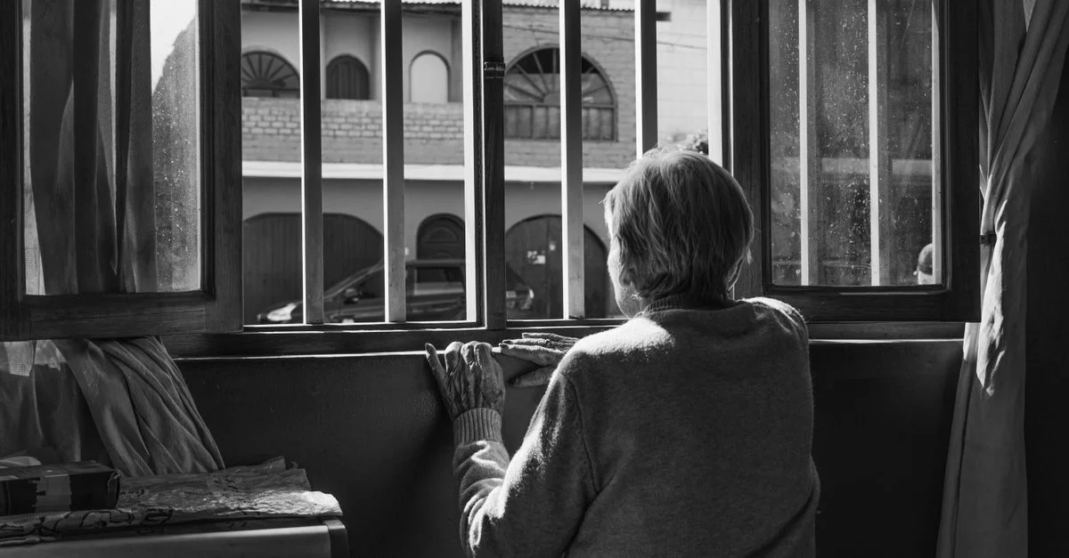Une femme senior regarde par une fenêtre, symbolisant la solitude et la contemplation.