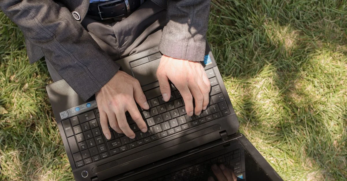 Homme travaillant sur un ordinateur portable en costume assis sur l'herbe, en extérieur pendant la journée.