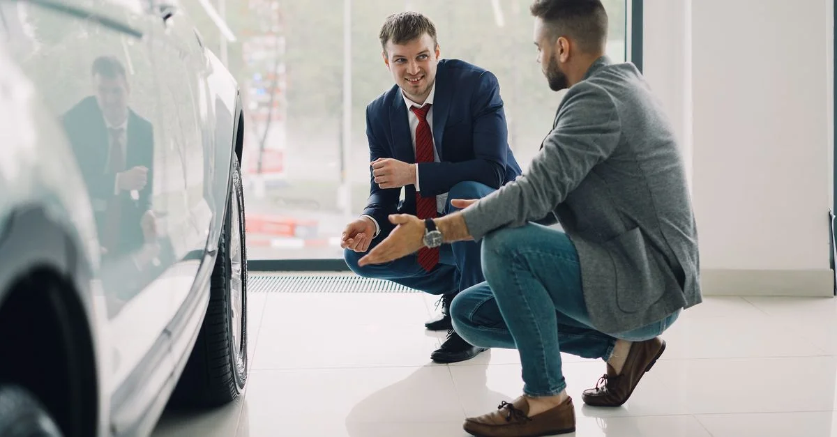 Deux hommes discutent des caractéristiques d'une voiture dans une salle d'exposition, agenouillés près d'un véhicule.
