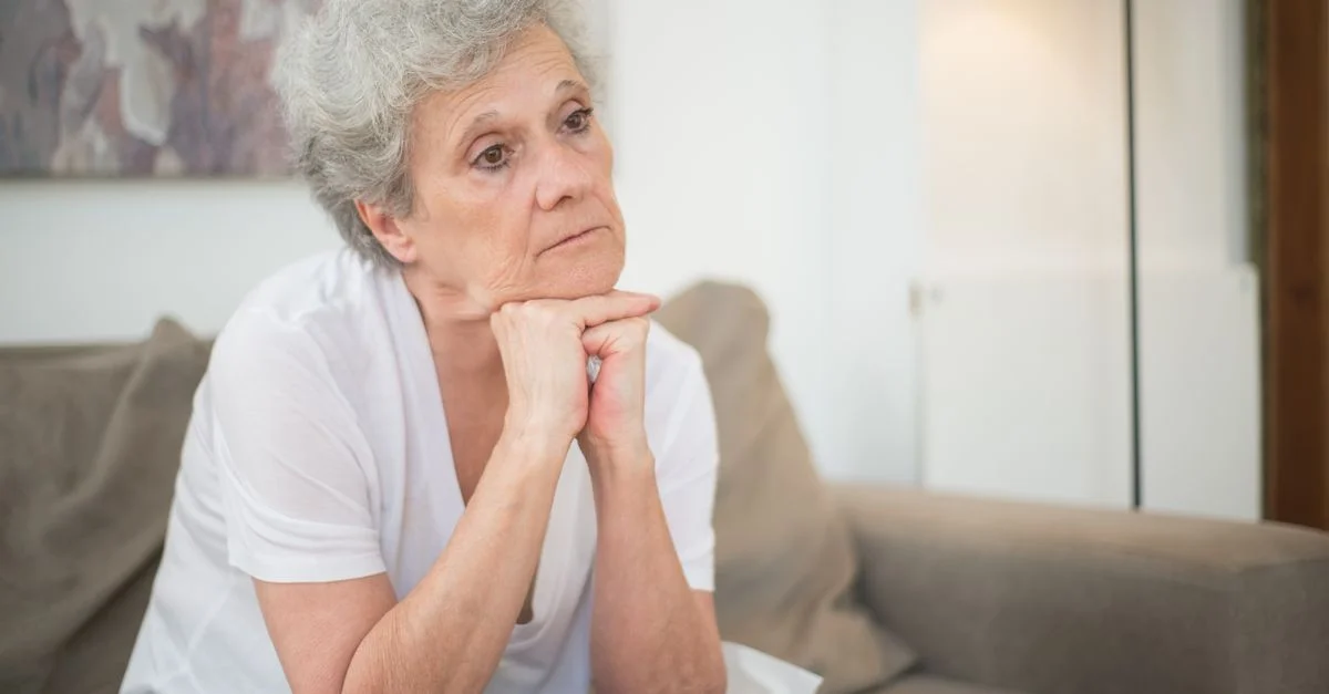 Une femme senior pensive avec des cheveux gris assise à l'intérieur, réfléchissant.