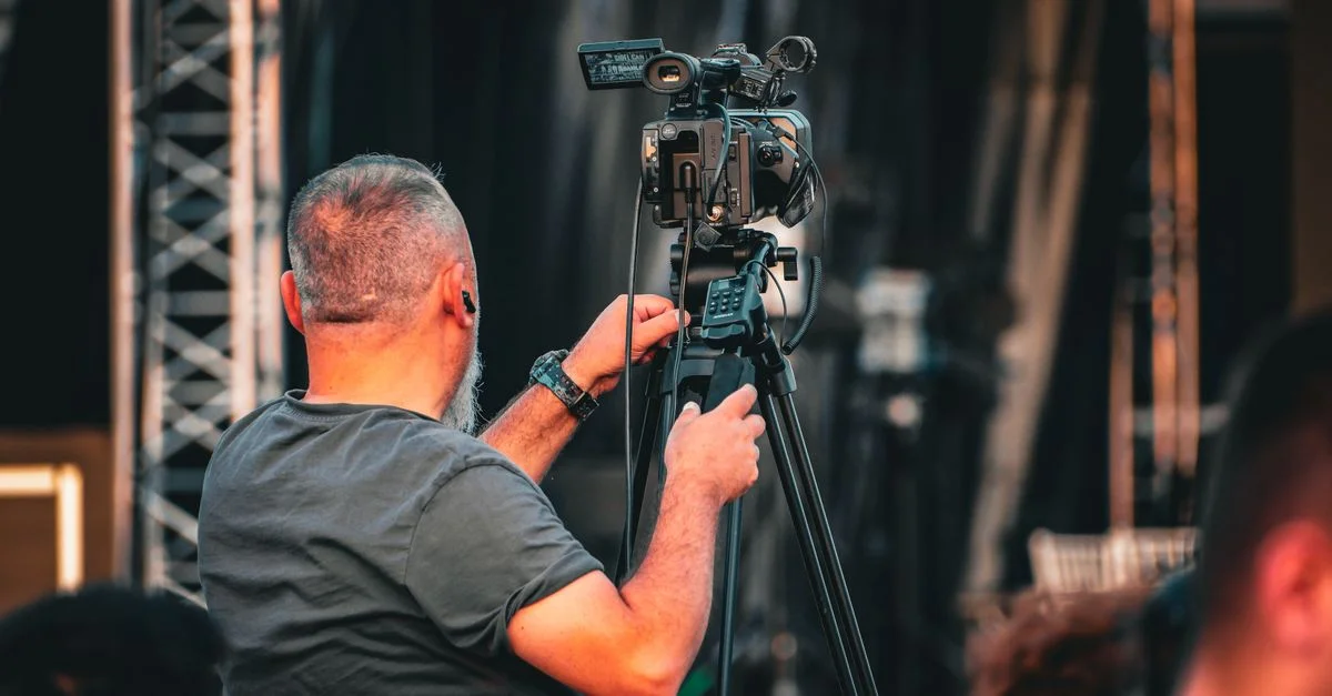Un caméraman professionnel utilisant une caméra vidéo sur trépied lors d'un événement en plein air, capturé de derrière.