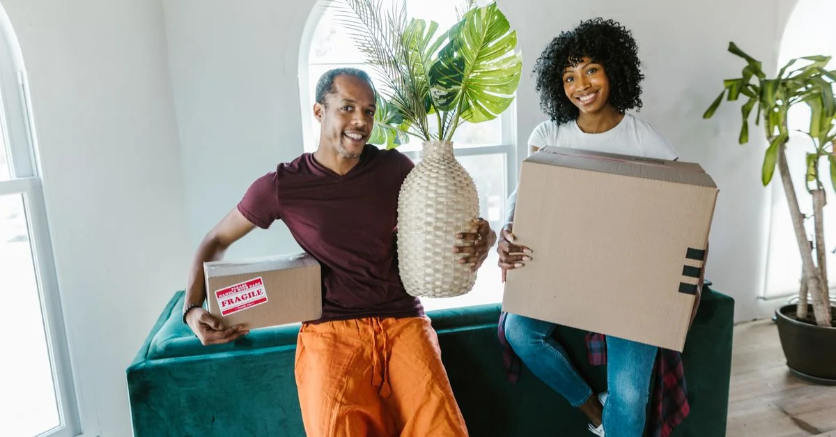 Couple souriant en emménageant dans une nouvelle maison, portant des cartons et une plante.