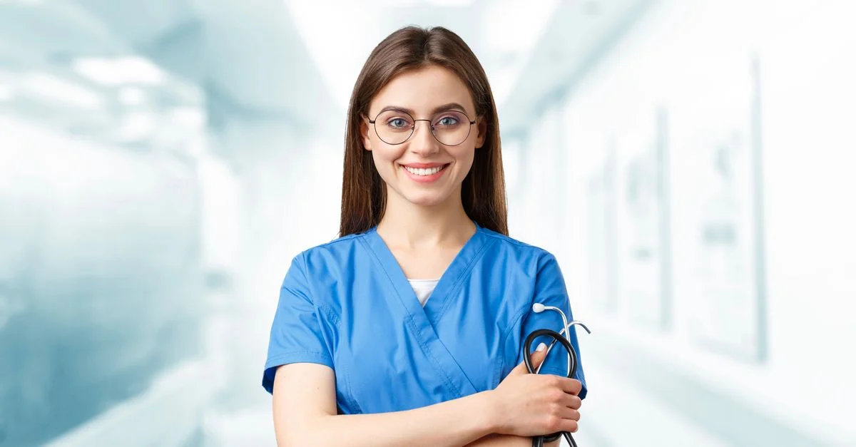 Jeune infirmière en blouse bleue souriante tenant un stéthoscope dans un couloir d'hôpital.