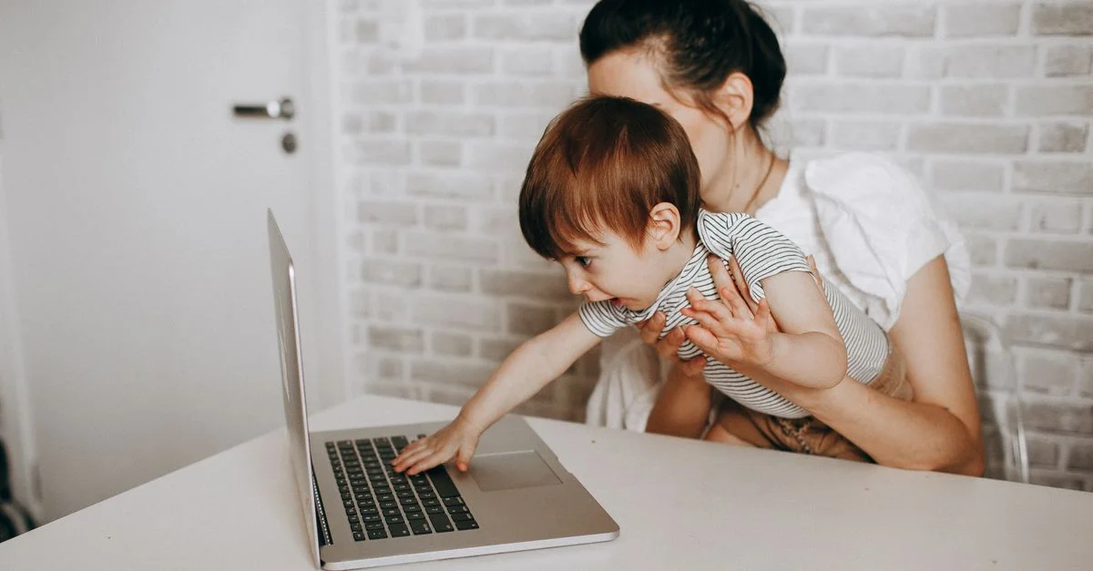 Une jeune mère tient son bébé dans ses bras tandis qu'elles utilisent toutes deux un ordinateur portable, illustrant ainsi la parentalité moderne.