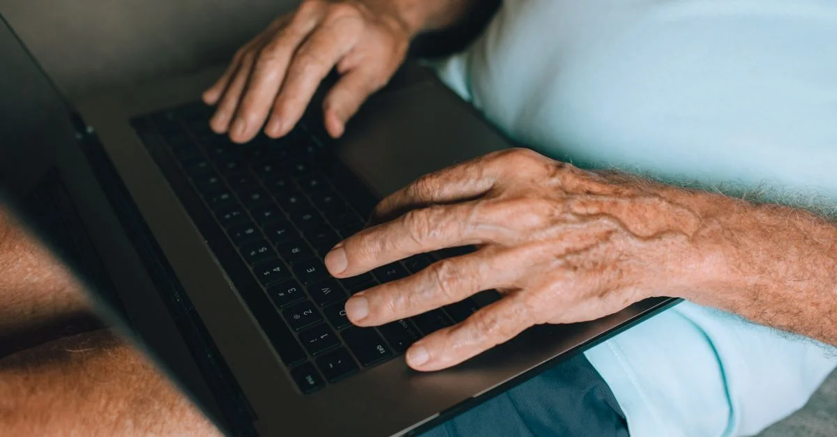 Gros plan sur les mains d'un homme senior tapant sur un clavier d'ordinateur portable à l'intérieur, mettant en avant l'utilisation de la technologie.