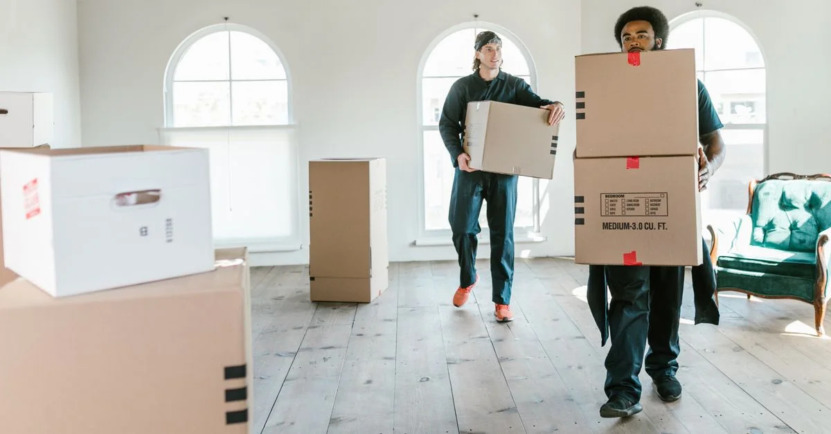 Deux déménageurs portant des boîtes dans une pièce ensoleillée, prêts à aider au déménagement.