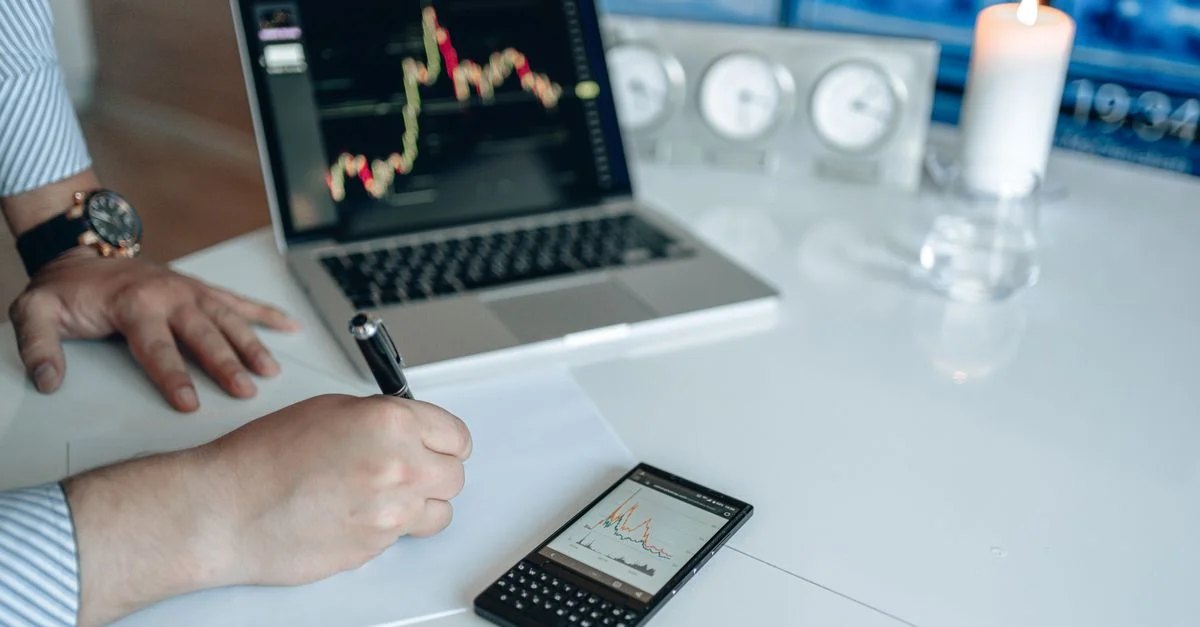 Analyste commercial écrivant avec un ordinateur portable et un smartphone affichant des graphiques de tendance sur un bureau blanc.