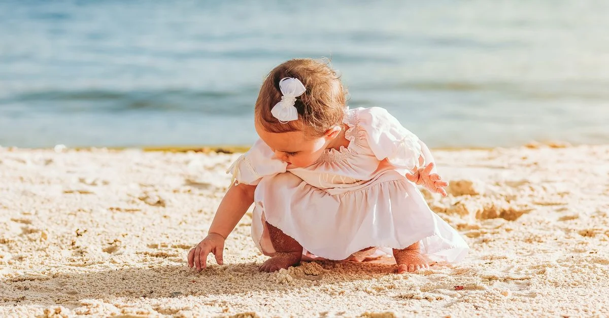 Charmante petite fille en robe blanche explorant le sable sur une plage ensoleillée.