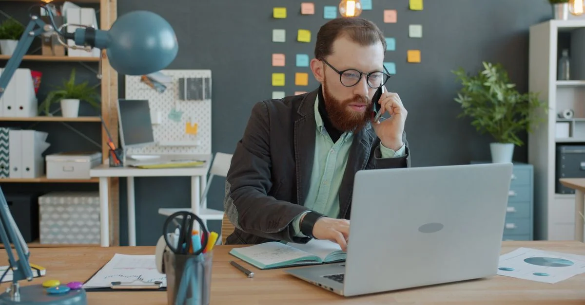 Homme d'affaires professionnel en pleine conversation téléphonique tout en travaillant sur son ordinateur portable dans un bureau moderne.
