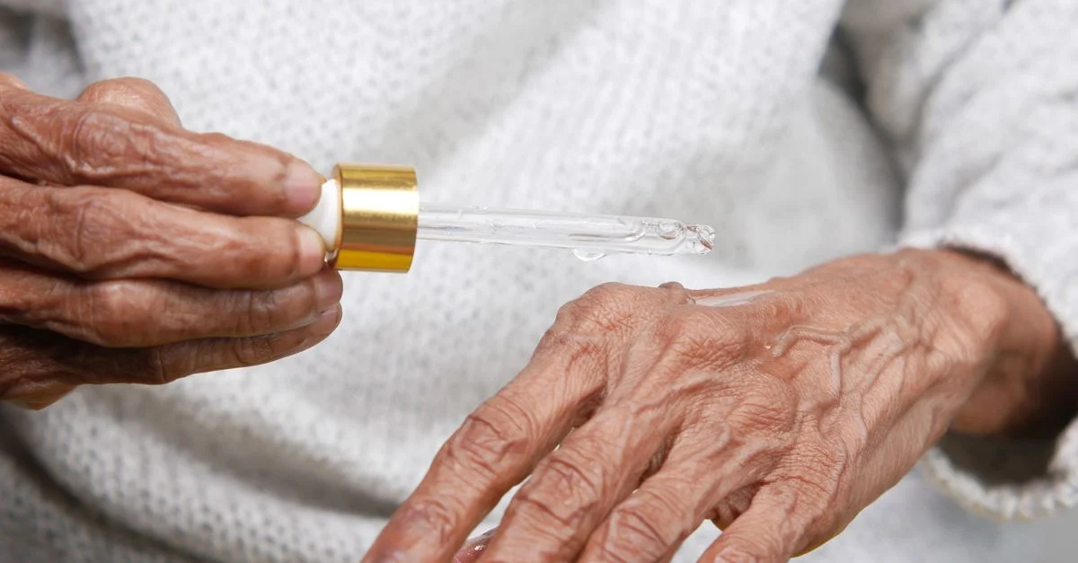 Gros plan sur des mains âgées appliquant un sérum avec un compte-gouttes, mettant l'accent sur les soins de la peau et le vieillissement.